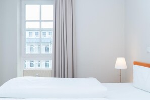 Ein Hotelzimmer mit großem Fenster, einem Bett, einem Nachttisch mit Lampe und Blick auf umliegende Gebäude.