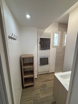 Un couloir étroit avec une étagère en bois, un radiateur sur lequel sont posées des serviettes et un évier blanc.