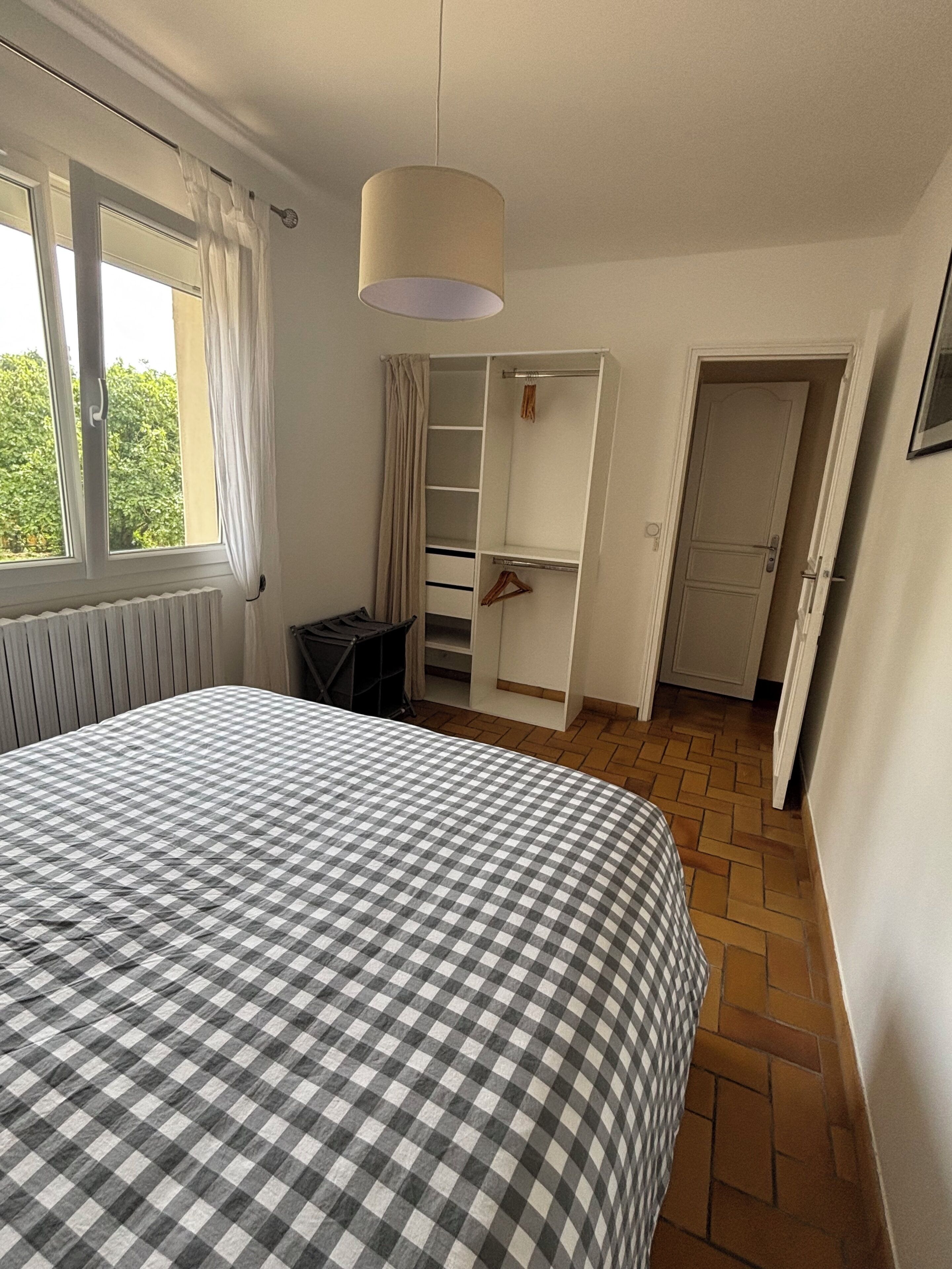 Un dormitorio con una colcha a cuadros, una ventana con cortinas, un radiador, un armario y una puerta que da a otra habitación.