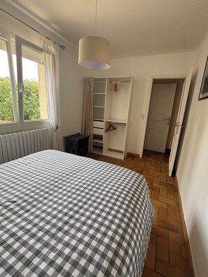 Une chambre avec une couvre-lit à carreaux, une fenêtre avec des rideaux, un radiateur, une armoire et une porte donnant sur une autre pièce.