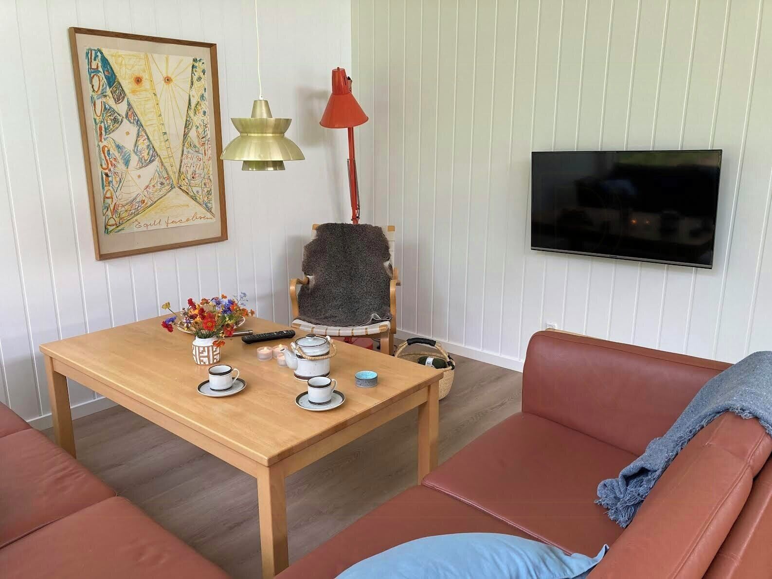 A living room with a wooden table, a sofa, a TV mounted on the wall, a chair, and a lamp.