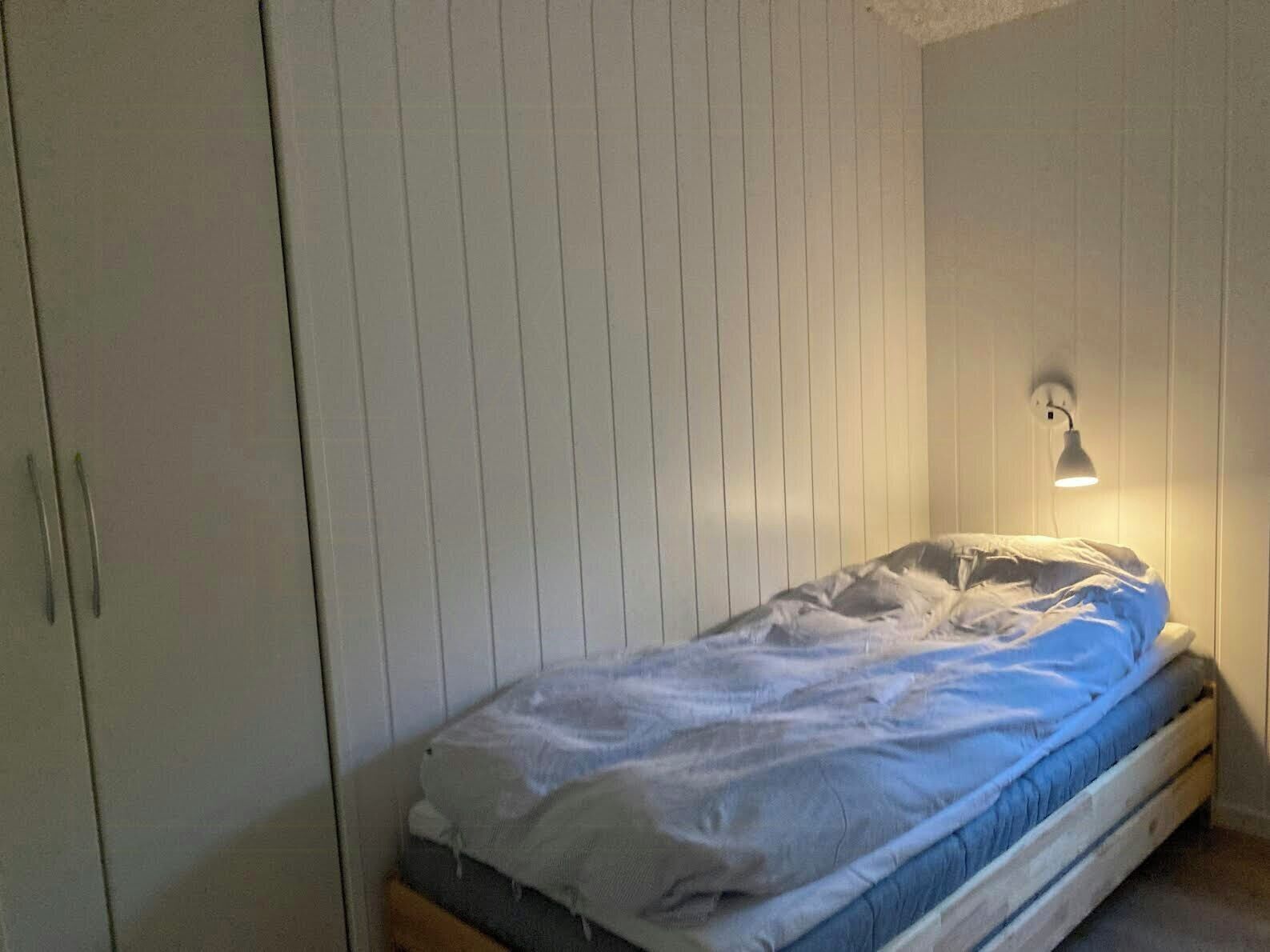 Ein Einzelbett mit weißer und blauer Matratze, eine Wandlampe und ein Kleiderschrank mit Griff.