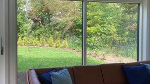 A modern living room with a brown leather sofa, a glass door leading to a garden with grass and trees. - Modern wooden house near Kollerup Forest (Fjettitslev)