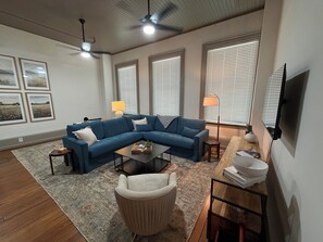 Ein Wohnzimmer mit einem blauen Ecksofa, einem kleinen runden Tisch, einem beigen Sessel und einem Couchtisch. Es gibt drei Fenster mit Jalousien, einen Deckenventilator und einen flachbildfernseher an der Wand.