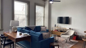 A modern living room with a blue sofa, a wooden coffee table, a TV mounted on the wall, and a ceiling fan. - Downtown Delight: Cozy + Chic 2 Bedroom Loft in Downtown Macon (Macon)