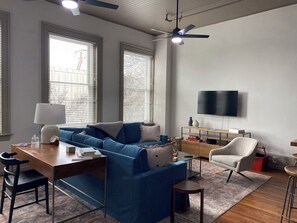 Ein modernes Wohnzimmer mit einem blauen Sofa, einem Holztisch, einem an der Wand befestigten Fernseher und einem Deckenventilator.