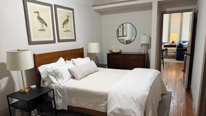 A neatly made bed with white linens, a bedside table with a lamp, and a framed bird illustration on the wall. - Downtown Delight: Cozy + Chic 2 Bedroom Loft in Downtown Macon (Macon)