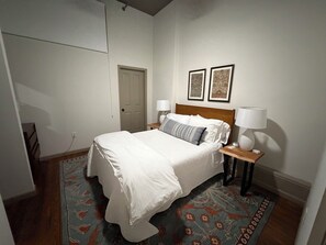 Una cama bien tendida con cabecera de madera, dos lámparas de noche y una alfombra estampada sobre piso de madera.