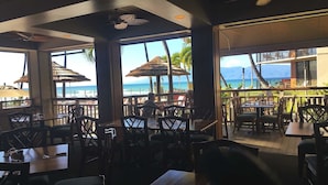 Un comedor interior con mesas y sillas, amplias ventanas que ofrecen vista a la playa y al océano, y sombrillas de paja.