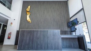 A reception area with a modern design, including a grey textured wall, a gold abstract sculpture, and a flat-screen TV. - Rbk Dpto GIM-3 personas-Lavanderia-WIFI (Santiago)