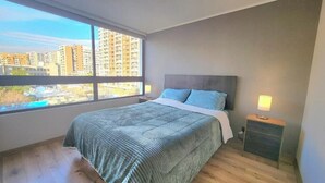A bedroom with a large window, a bed with a grey headboard, a bedside table with a lamp, and a view of buildings and a pool outside. - Rbk Dpto GIM-3 personas-Lavanderia-WIFI (Santiago)