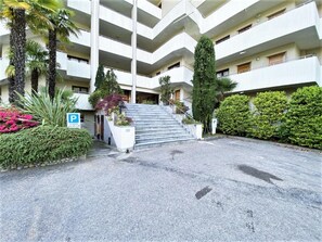 Edifício de vários andares com área de estacionamento, escadas que levam à entrada e vegetação ao redor.