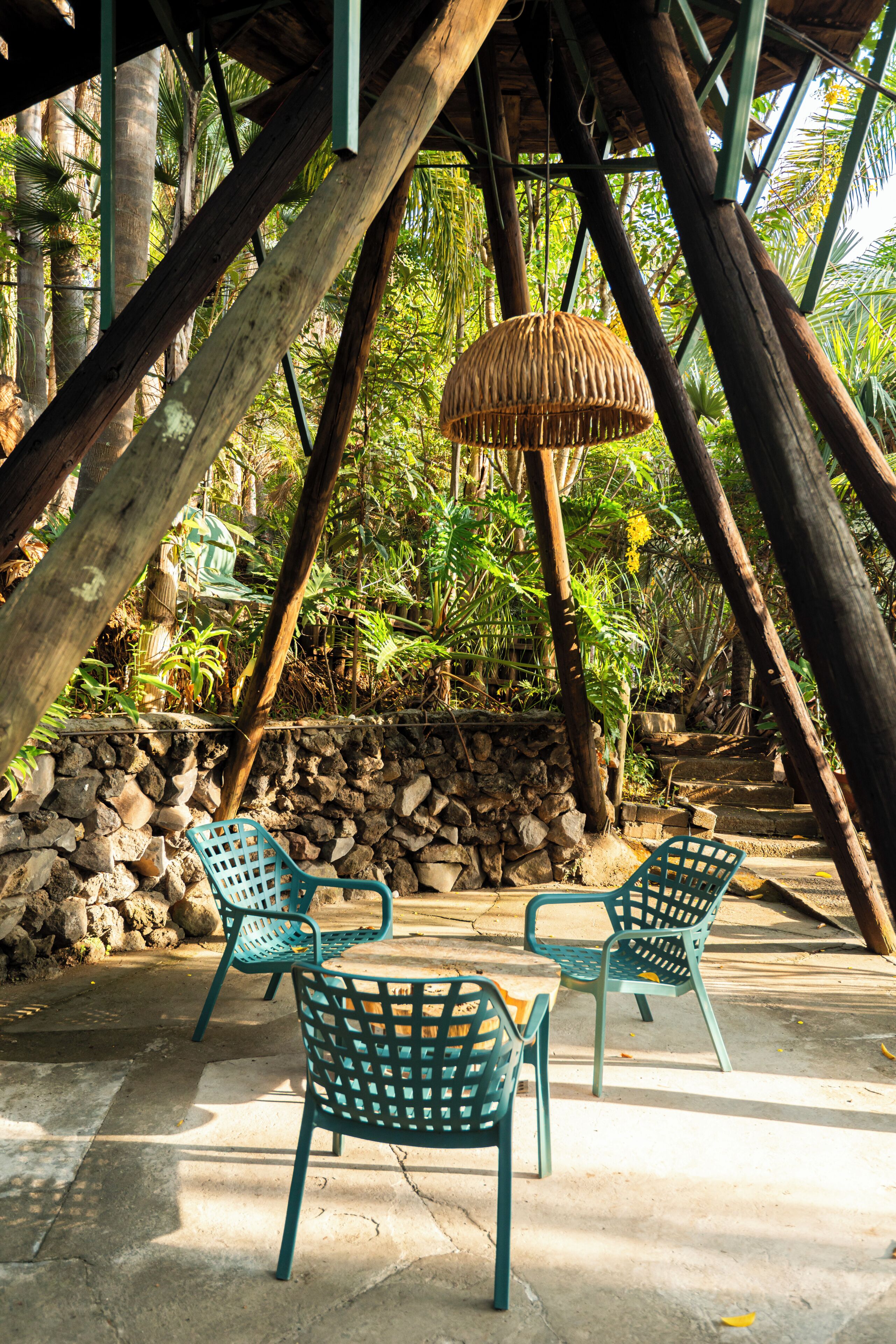 A covered outdoor seating area with chairs and a woven lamp, surrounded by stone walls and tropical vegetation.