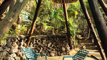 A covered outdoor seating area with chairs and a woven lamp, surrounded by stone walls and tropical vegetation.