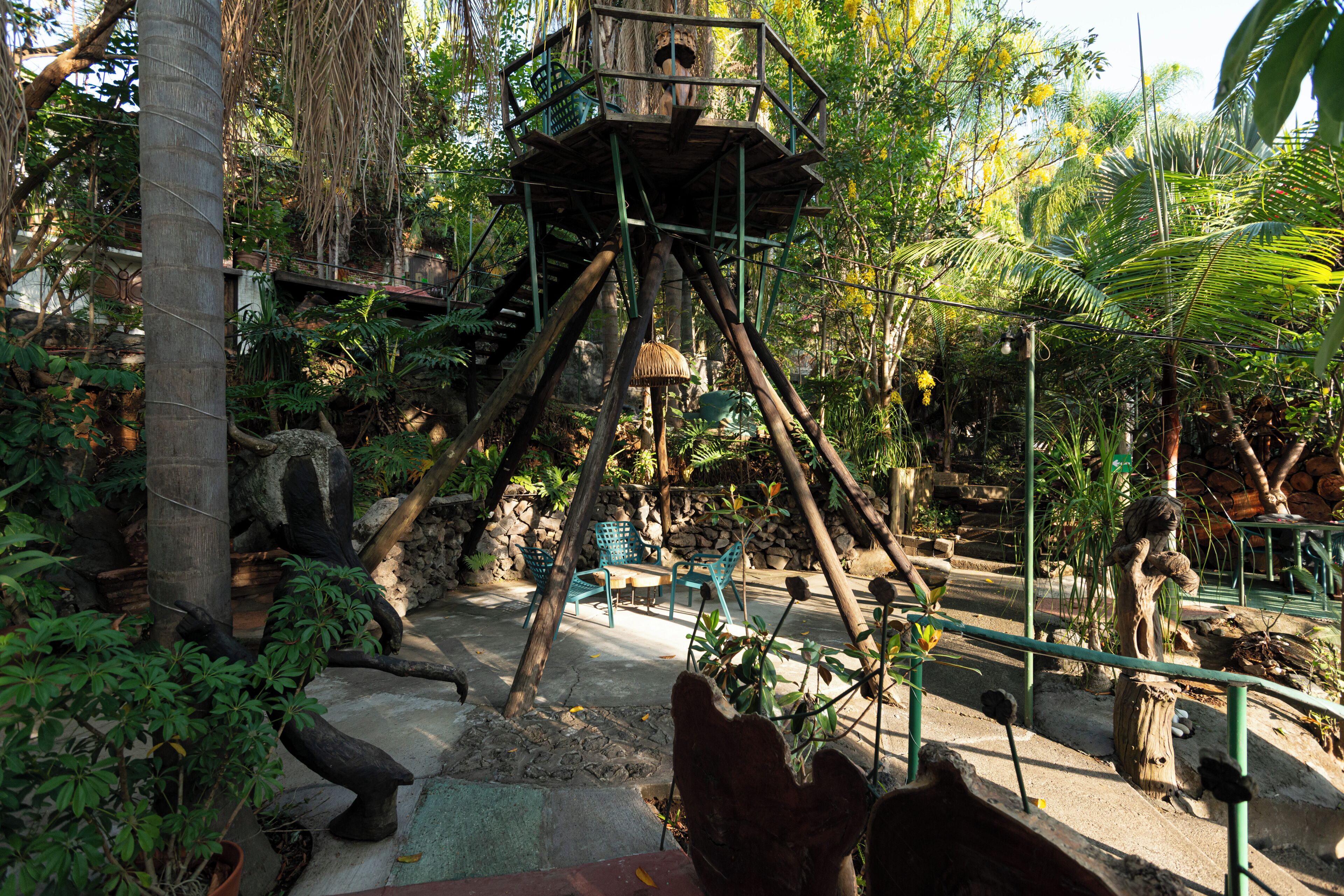 Una torre di osservazione in legno con una piattaforma, circondata da vegetazione tropicale e un muro in pietra.