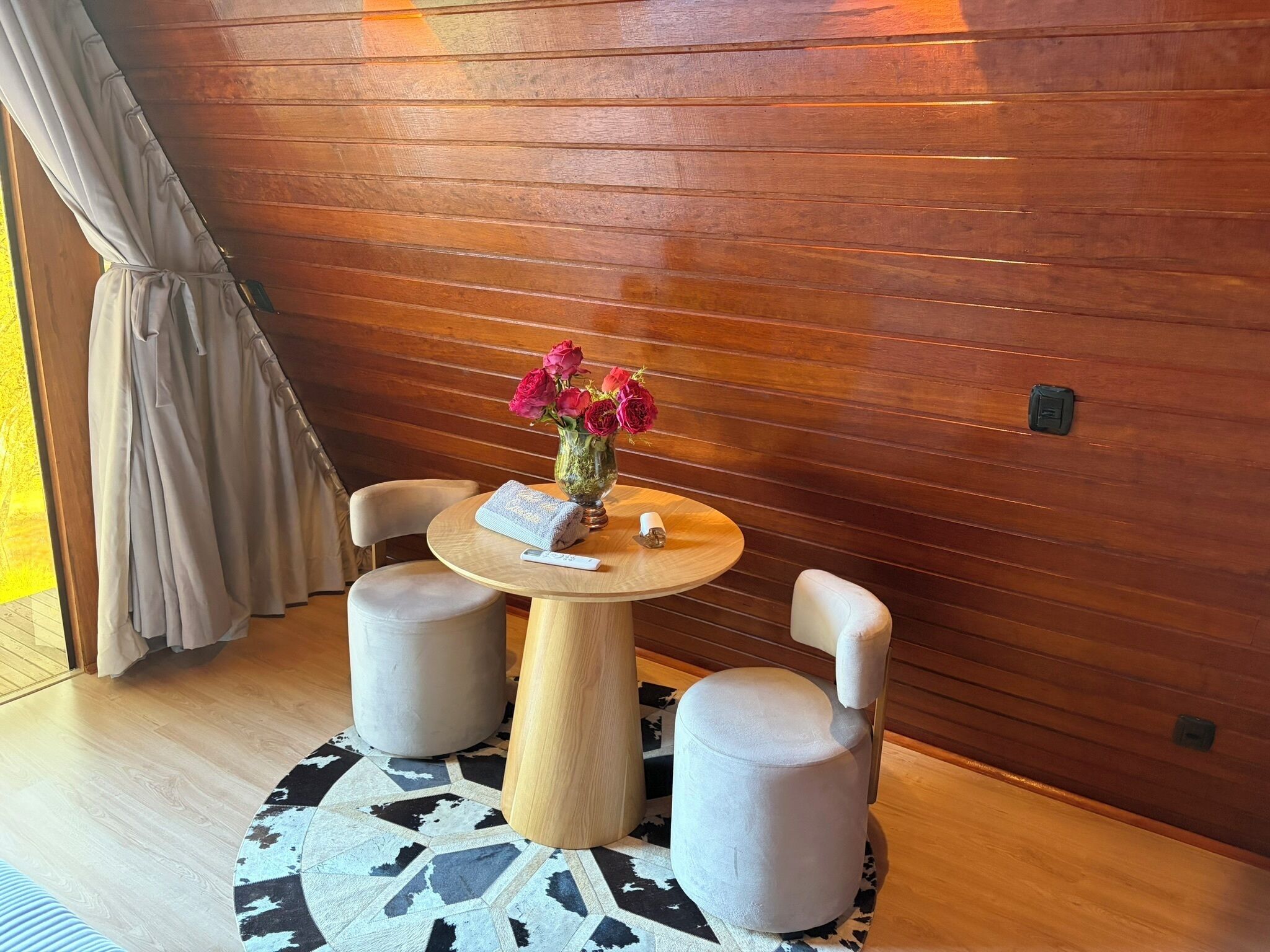 A room with a wooden paneled wall, a round wooden table with two chairs, a vase of flowers, and a remote control on the table.