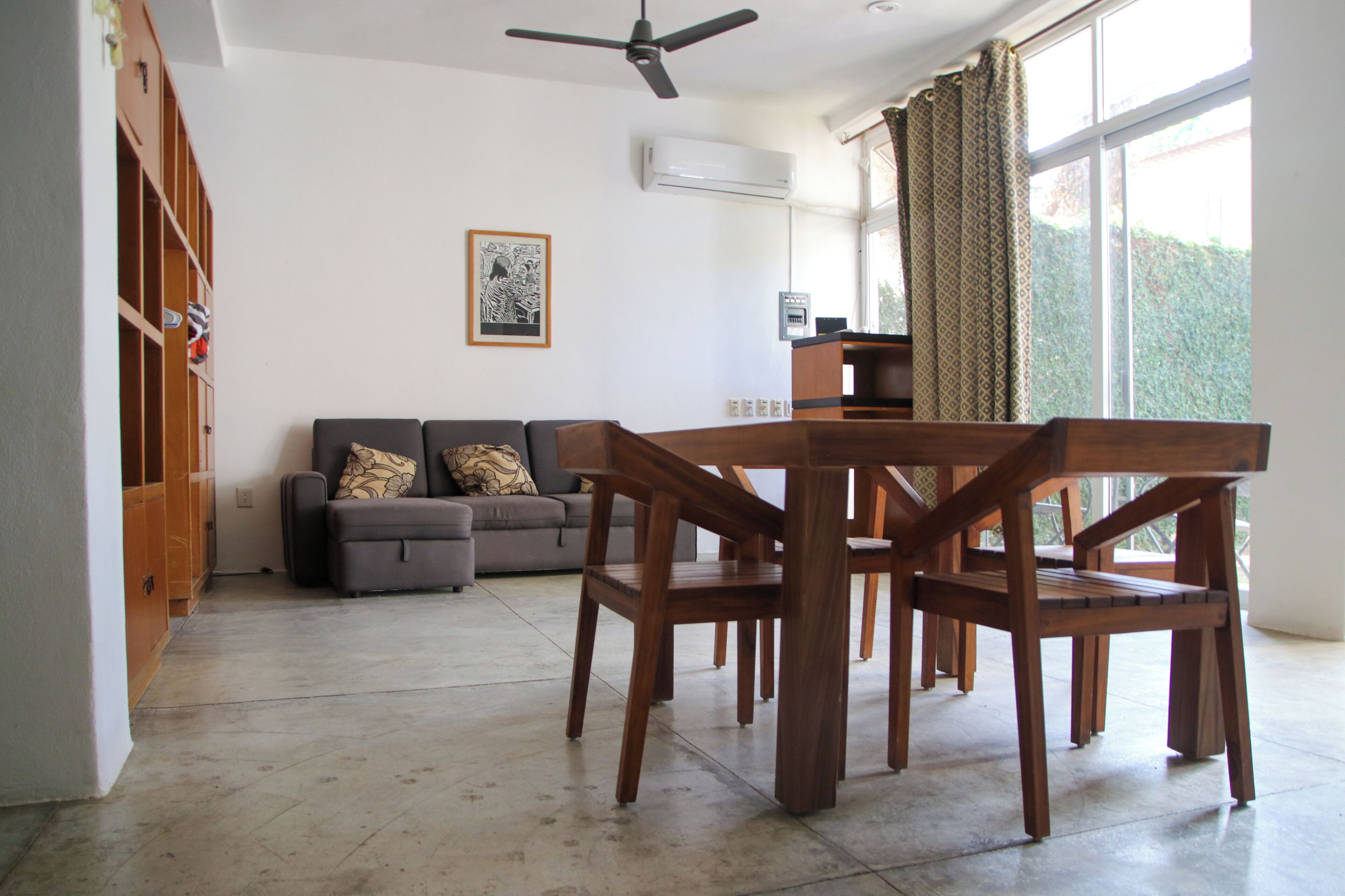 Uma sala de estar com sofá, mesa de jantar de madeira, cadeiras, ventilador de teto e janela com cortinas.