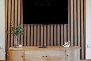 A wooden TV stand with a flat-screen TV mounted above it, a vase with green plants, a remote control, and a heart-shaped decoration. - Stylish 2BR 2BA | Free Parking Near Bluewater (Dartford)