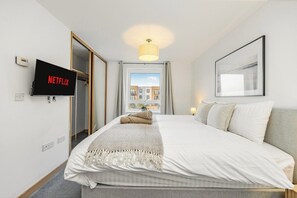A hotel room with a bed, a television displaying 'NETFLIX,' a window with a view of buildings, and a framed picture on the wall. - Stylish 2BR 2BA | Free Parking Near Bluewater (Dartford)