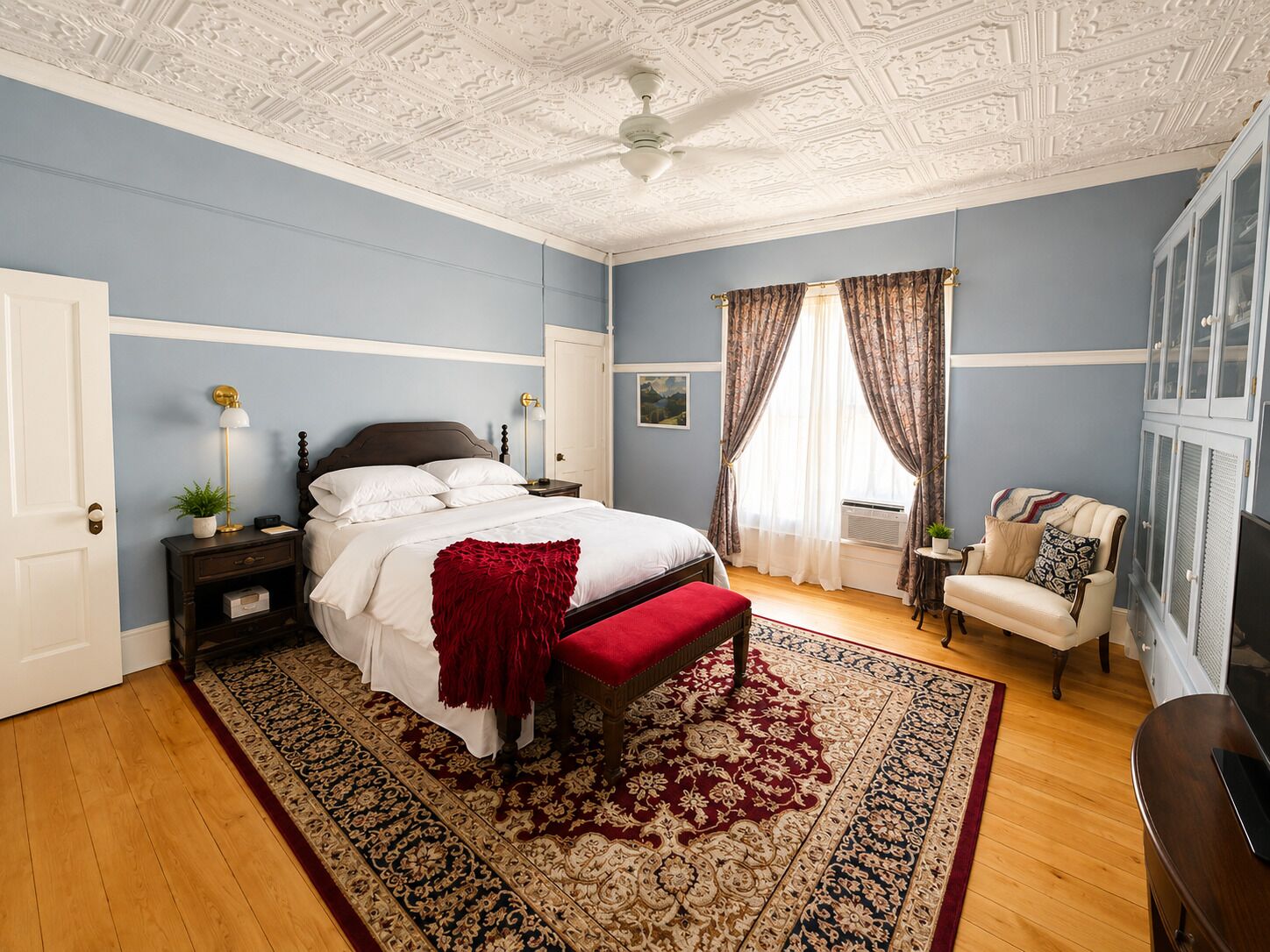 A bedroom with a large bed, a red throw blanket, a wooden bench, a chair, a nightstand, a lamp, and a ceiling fan.