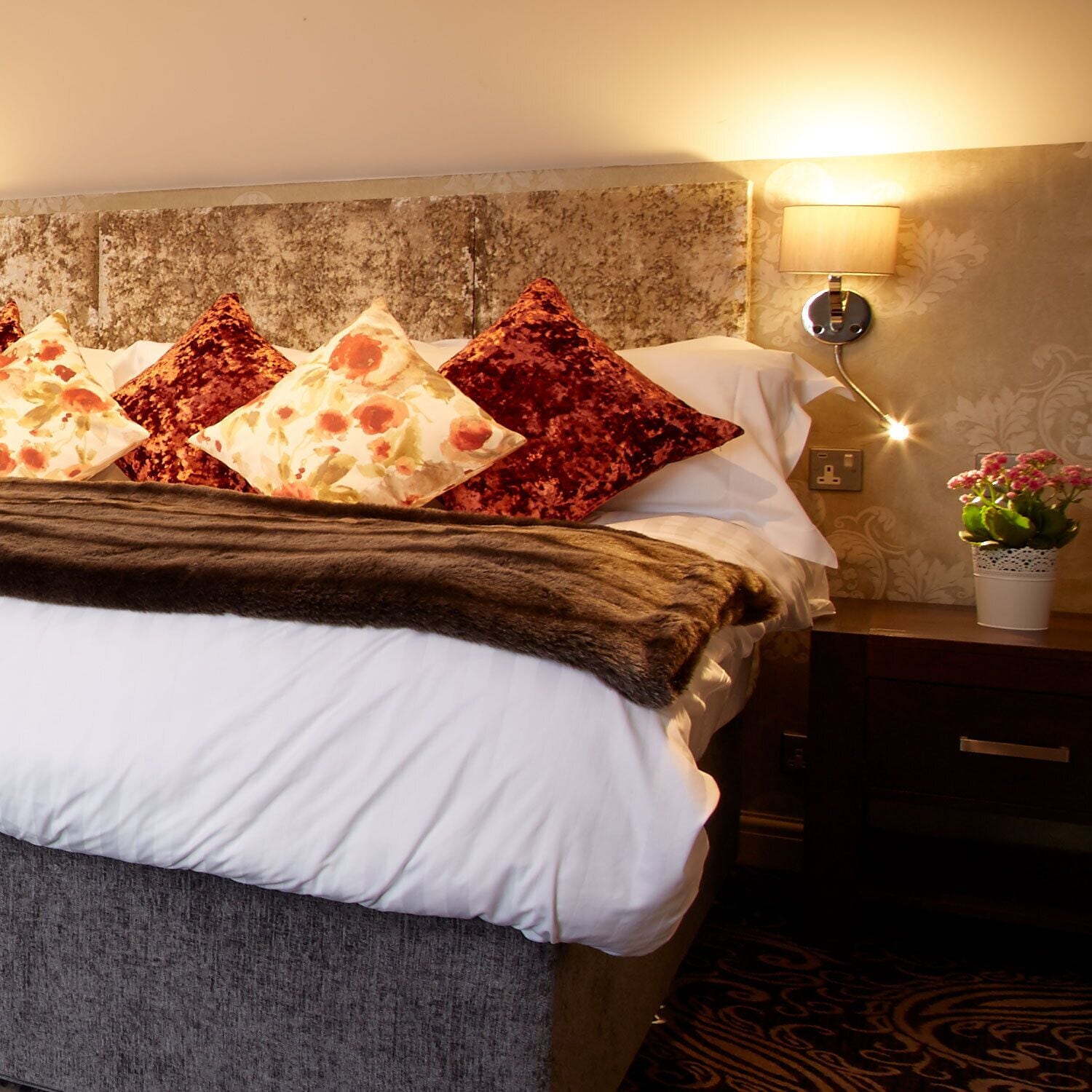 A hotel room with a bed, pillows, a nightstand with a lamp, and a potted plant.