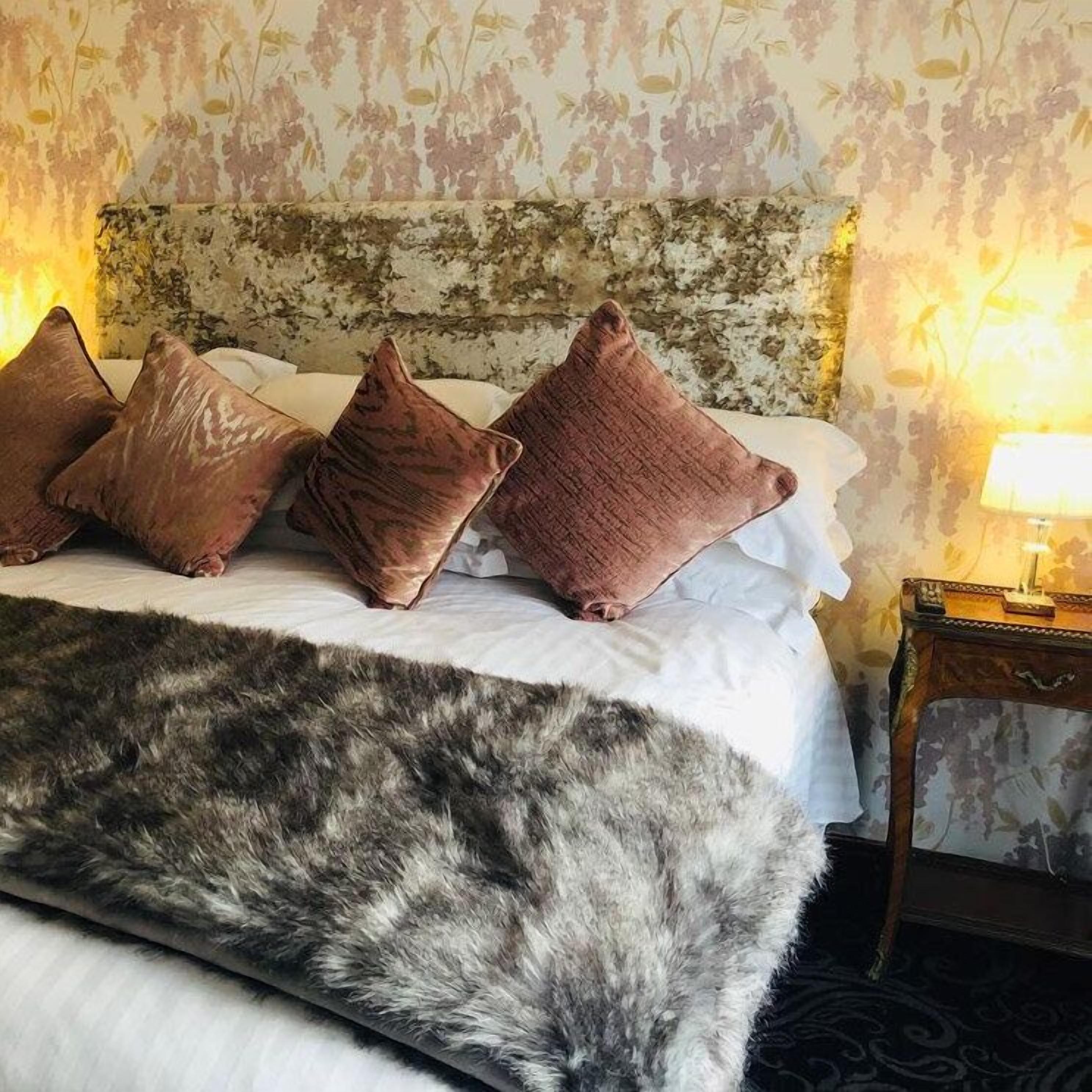 Una cama con cabecera blanca, varios almohadones, un plaid de piel y una lámpara en la mesita de noche.