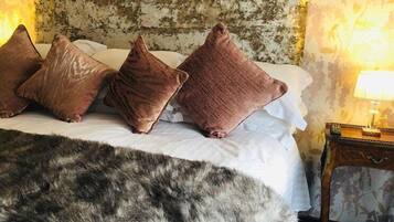 Una cama con cabecera blanca, varios almohadones, un plaid de piel y una lámpara en la mesita de noche.