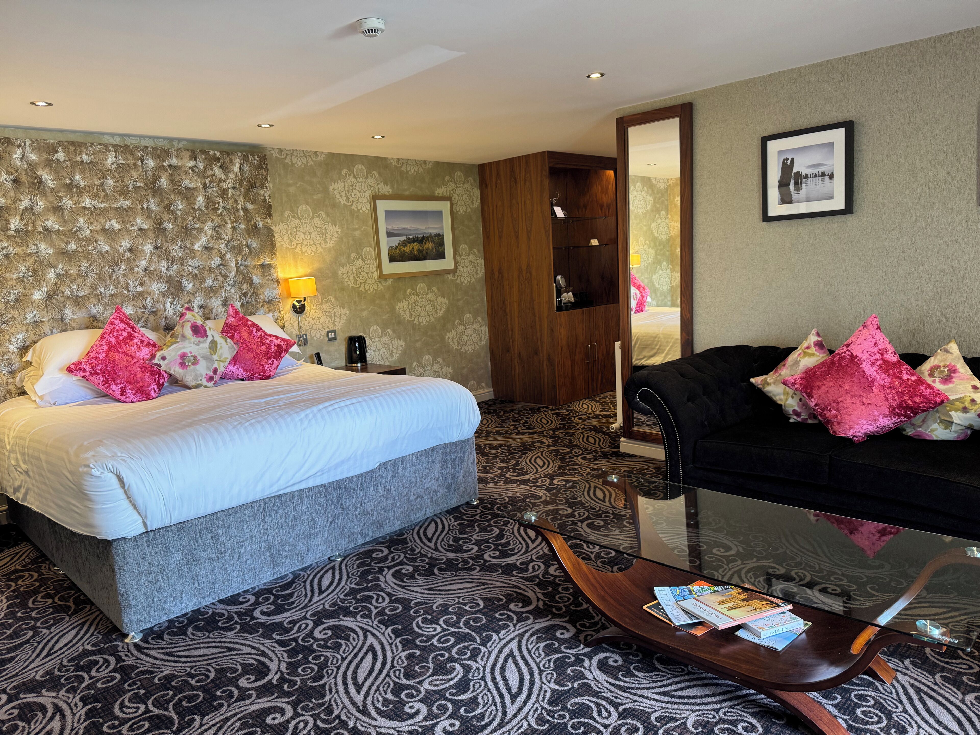 A hotel room with a bed, a sofa, a glass coffee table, and a patterned carpet.