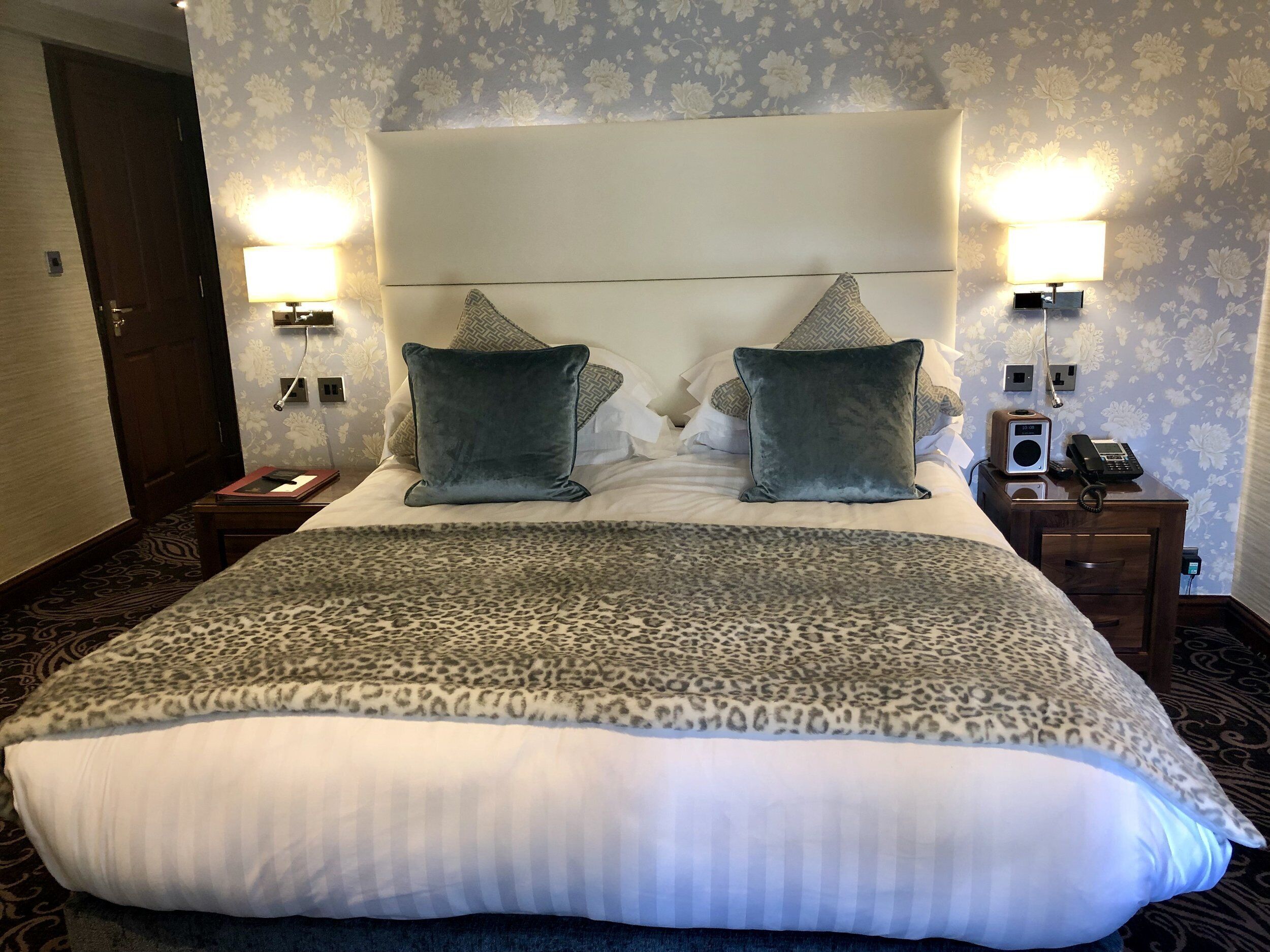A hotel room with a large bed, two bedside lamps, a nightstand with a phone and a radio, and floral wallpaper.