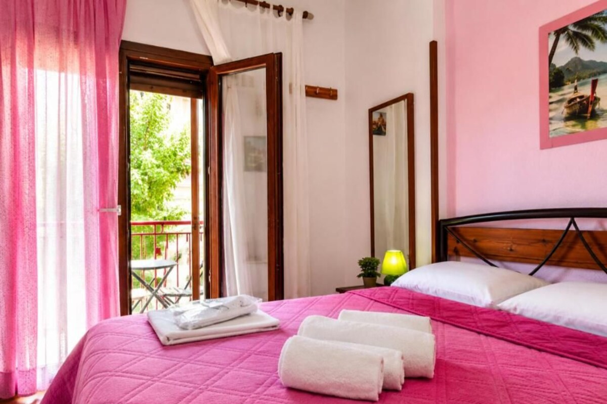 Un dormitorio con cama, almohadas, una mesita de noche con lámpara, una ventana con cortinas y un balcón con silla.