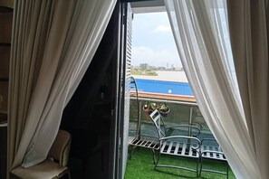Ein Zimmer mit Blick auf ein Gewässer und ein Gebäude mit blauem Dach. Draußen stehen ein Sessel und ein Tisch mit Stühlen.