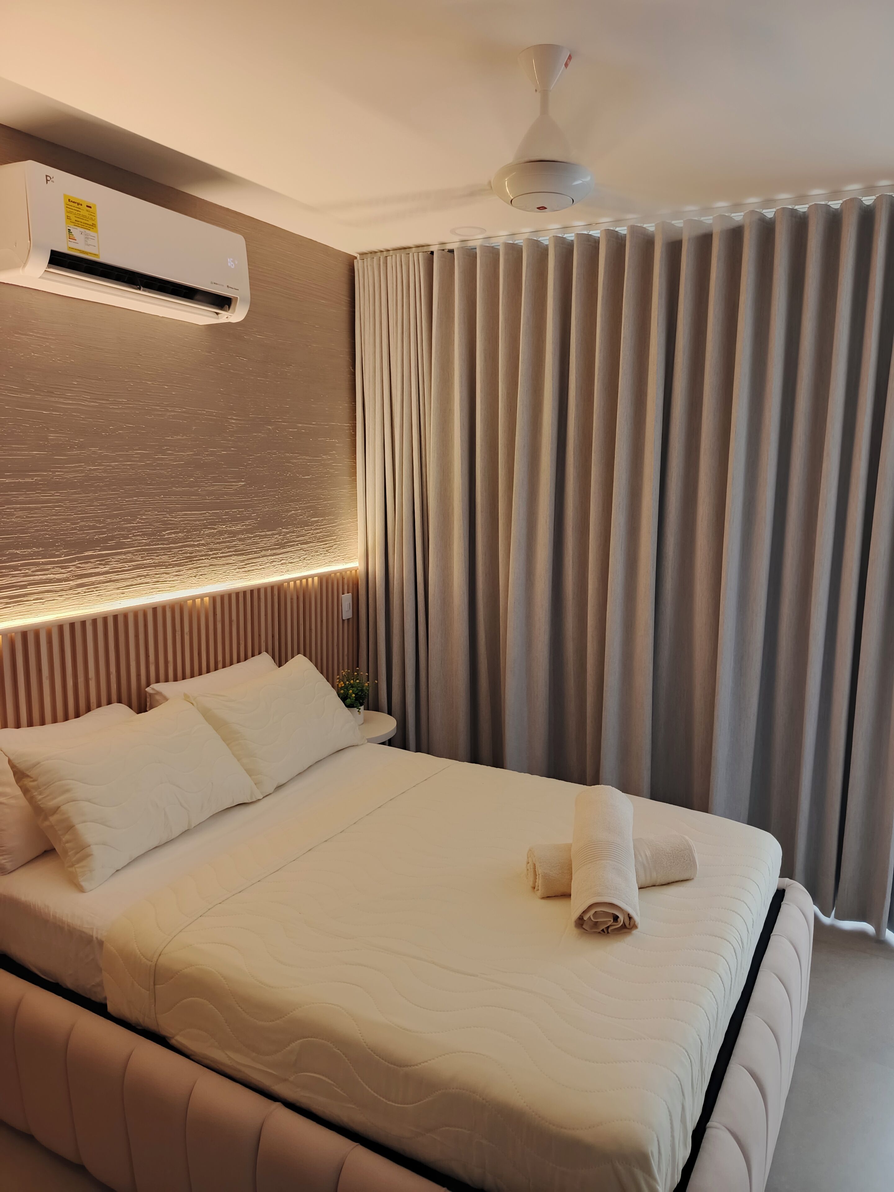 A hotel room with a bed, a wall-mounted air conditioner, a ceiling fan, and a window with curtains.