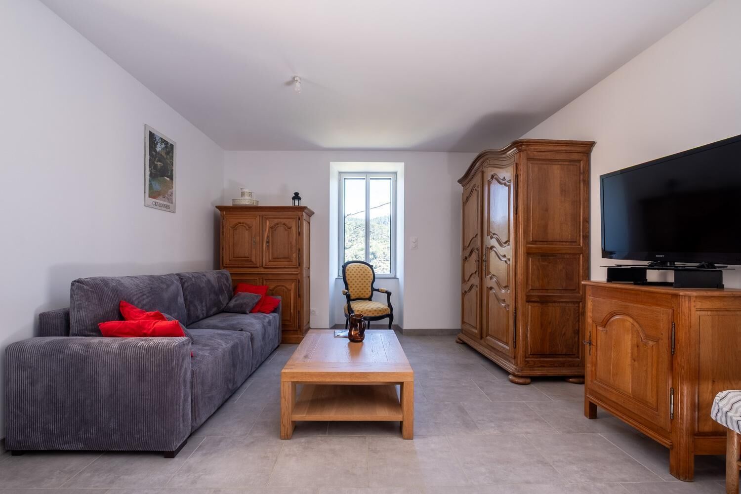 Un salon avec un canapé gris, une table basse en bois, un téléviseur posé sur un meuble en bois et une fenêtre laissant entrer la lumière naturelle.
