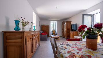 Une pièce spacieuse avec un meuble en bois, un canapé, une table à manger recouverte d’une nappe à motifs floraux et un téléviseur posé sur un meuble en bois.