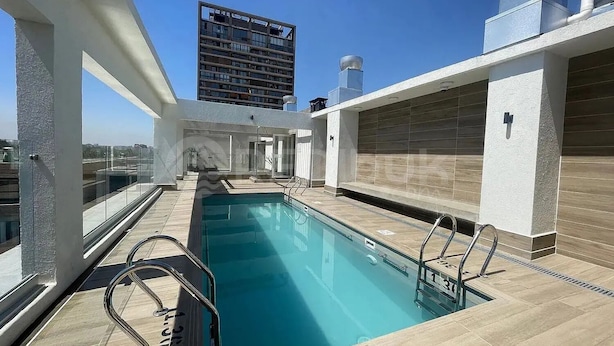 Piscina no terraço com guarda-corpos de vidro transparente, um edifício moderno ao fundo e céu claro.