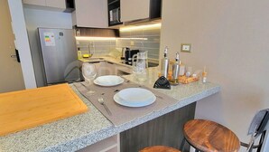 Cozinha compacta com geladeira, micro-ondas e pia. Há dois bancos e uma tábua de corte de madeira no balcão.