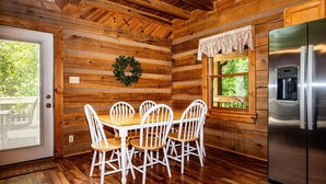 A wooden cabin interior with a dining table, chairs, a refrigerator, and a window with a view of greenery. - Snuggled Inn | Private Brown County Log Cabin on 12 Wooded Acres (Bloomington)