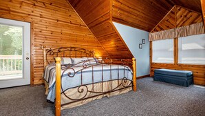 A bedroom with a wooden ceiling, a bed with a metal frame, a bench, and a window with blinds. - Snuggled Inn | Private Brown County Log Cabin on 12 Wooded Acres (Bloomington)