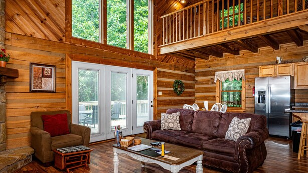 A cabin interior with a wooden floor, a brown leather sofa, a white coffee table, and a kitchen area with a stainless steel refrigerator. - Snuggled Inn | Private Brown County Log Cabin on 12 Wooded Acres (Bloomington)