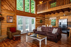 A cabin interior with a wooden floor, a brown leather sofa, a white coffee table, and a kitchen area with a stainless steel refrigerator. - Snuggled Inn | Private Brown County Log Cabin on 12 Wooded Acres (Bloomington)