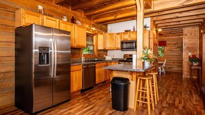 A wooden cabin interior with a stainless steel refrigerator, wooden cabinets, a microwave, and a bar area with stools. - Snuggled Inn | Private Brown County Log Cabin on 12 Wooded Acres (Bloomington)