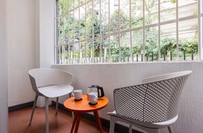 Ein Balkon mit zwei Stühlen, einem kleinen runden Tisch und einem Fenster mit Blick auf Grünflächen und Gebäude.