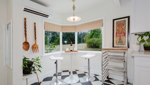 Une cuisine avec un carrelage à motifs, deux tabourets de bar blancs, un étagère en métal et une fenêtre donnant sur un paysage verdoyant.