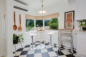 A kitchen with a checkered floor, two white bar stools, a metal rack, and a window with a view of greenery.