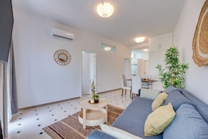 A modern living room with a sofa, a small round table with a plant, a dining area, and a kitchen in the background. - LXR Parador (Reformatted) 1 Bedroom (Benalmádena)