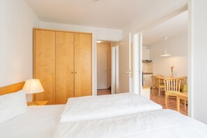 A hotel room with a bed, a wooden wardrobe, a dining area with chairs and a table, and a kitchen in the background. - Lichtdurchflutete Ferienwohnung mit Balkon in Sudostlage in Sellin (Sellin)