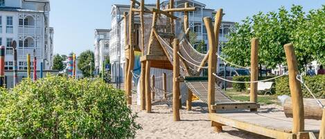 Un terrain de jeux avec un toboggan en bois et des structures d’escalade, entouré de sable et de verdure, avec des bâtiments en arrière-plan.
