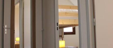 A room with a wooden door, a visible kitchen area with a window, and a bunk bed in the background.