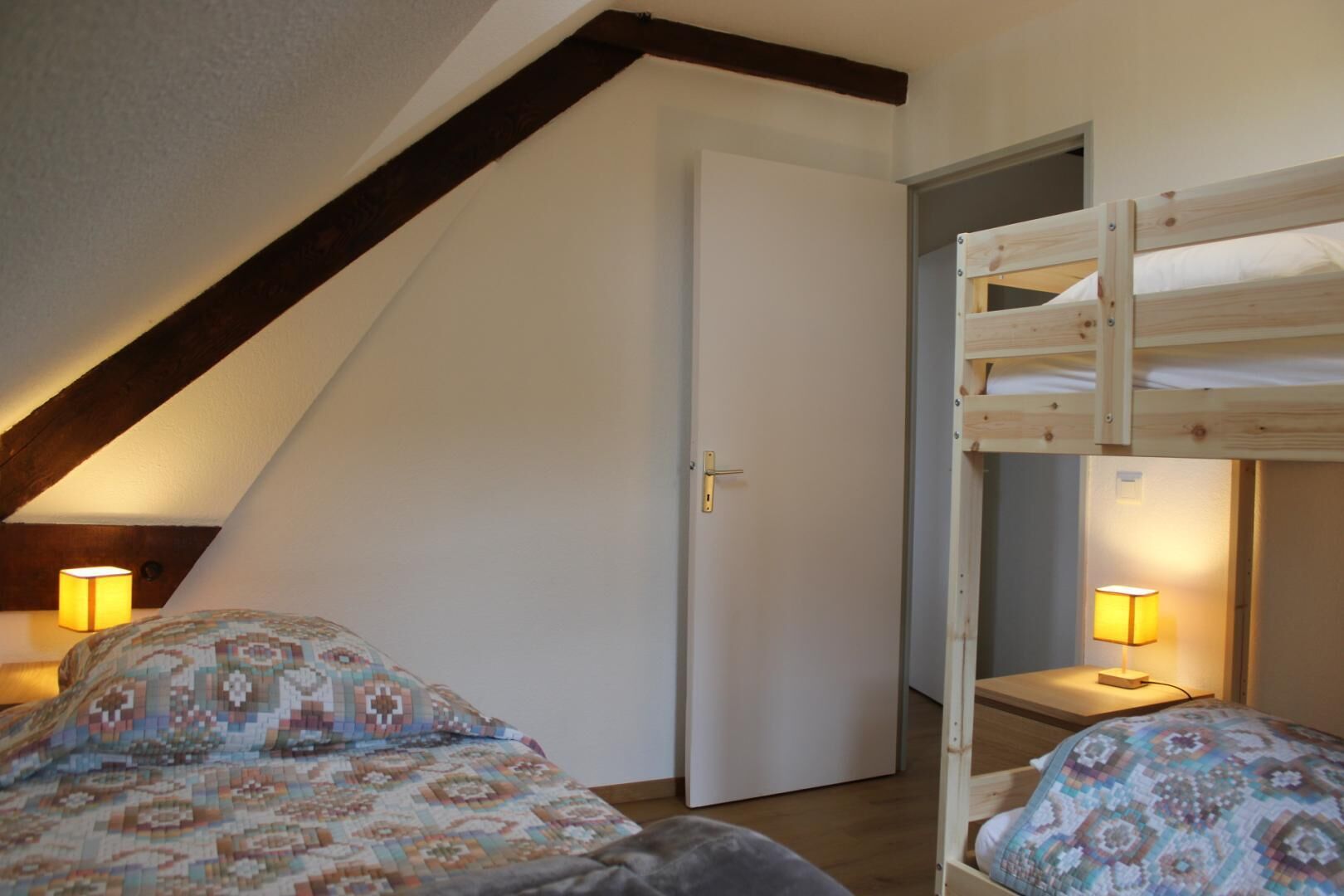 Een slaapkamer met een schuine kap, een stapelbed, twee nachtkastjes met lampen en een deur.