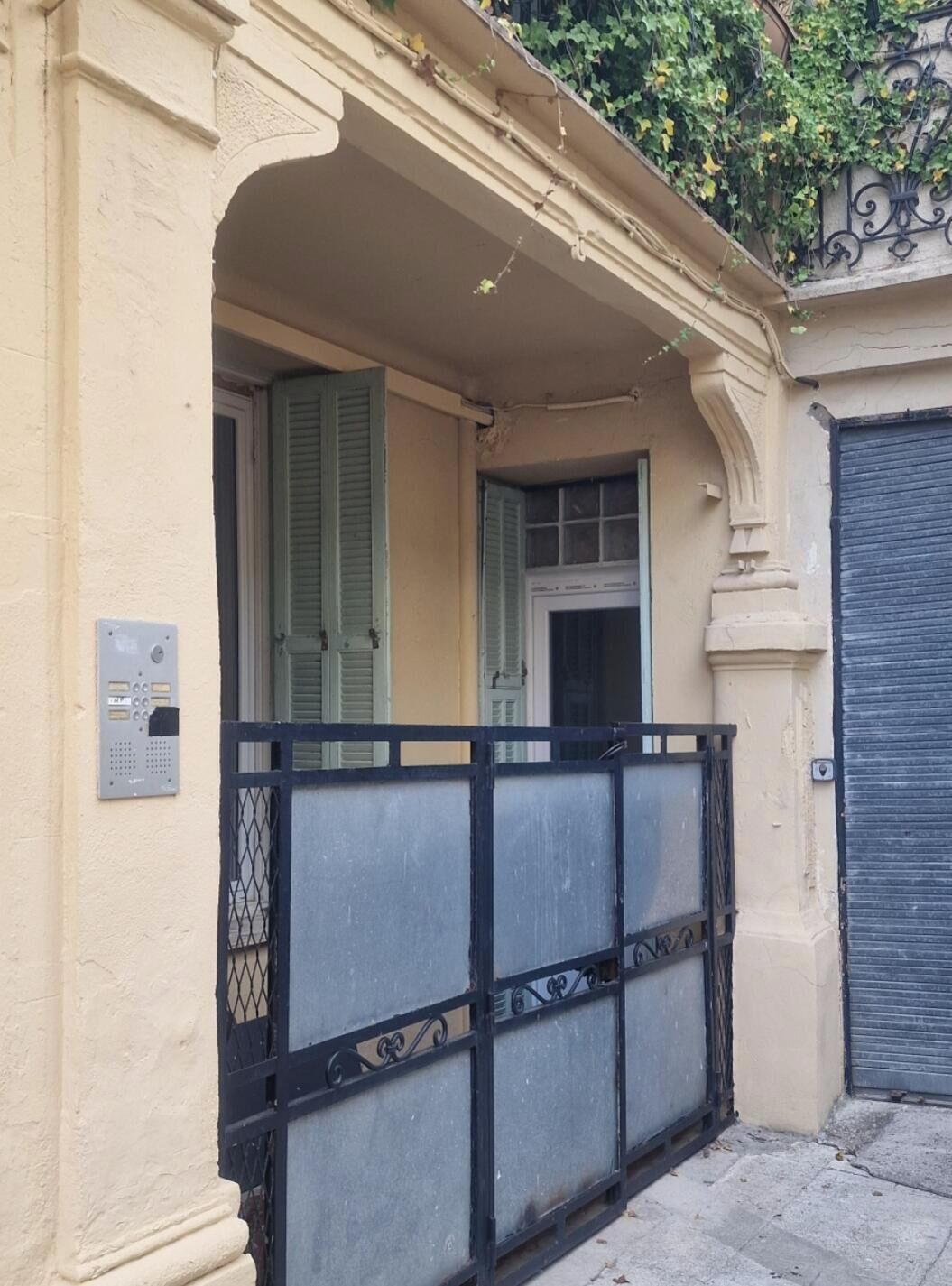 Fachada de prédio com portão fechado, porta com persiana e caixa de eletricidade visível.